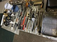 Photo showing wide assortment of kitchen utensils including spoons, peelers, spatulas, knives, tongs, measuring cups, mixing bowls, garlic press, ice cream scoop, mandoline, and drawer organizers arranged on floor.