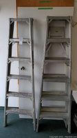 Two silver six-foot folding metal ladders shown side by side in a room against a white door.