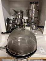 Stainless steel pots and pans stacked with mesh food cover dome and kitchen accessories in front.