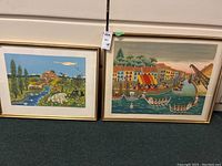 Photo showing two framed prints side by side on the floor. Left print shows pastoral village and river scene; right print shows boat harbor scene.