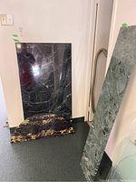 Overview of three marble table tops leaning against wall; includes large black marble, smaller black marble with gold veining, and tall green marble slab.