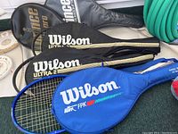 Four tennis racquets and their cases; Wilson Ultra FPK blue racquet and case, Wilson Ultra 2 Largehead black racquets, and two Prince racquets with black cases.