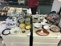 Overall view of all items in the lot arranged on two white dressers, showing tureen, plates, bowls, and decorative plate