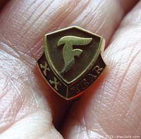 Close-up of the front side of the 14K yellow gold Firestone 20 years service pin showing the shield shape, embossed F letter, and Roman numerals XX with 'YEARS' inscription on the base.