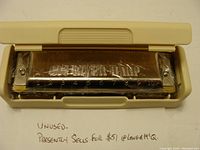 HOHNER Big River Harp harmonica inside a plastic protective case. Writing indicates the item is unused and currently retails at $51 at Lange & McQ.