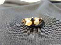 Close-up photo of ring showing front view with two pearls and blue stones on gold band.
