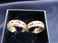 Pair of gold hoop earrings with detailed cut-out and textured design inside black jewelry box against dark fabric background.