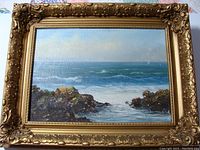 Frontal photo of the seascape painting featuring ocean waves between rocks, framed in an ornate gold frame with floral and scrollwork decoration.