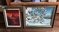 Photo showing both framed landscape paintings side by side on the floor; paintings show red sunset mountain scene and snowy pine tree cabin scene.