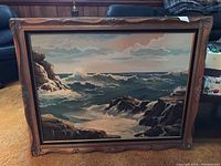 Full view of the framed seascape painting showing ocean waves crashing on rocks under a cloudy sky in a dark carved wooden frame.