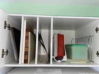 White cabinet interior with divided storage holding wood cutting boards, plastic cutting boards, metal rack, and Tupperware containers with green lids.