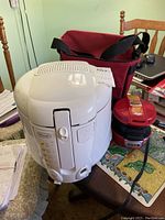 White DeLonghi deep fryer D680-C with temperature control and front handle, red Hamilton Beach sandwich maker, and maroon cooler bag on table.