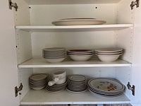 Open cupboard showing stacks of plates, bowls, smaller dishes, and some cups with Trellis Blossom floral pattern stoneware dishes.