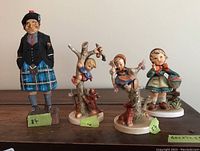 Group photo of one wooden Scottish man figurine and four Hummel ceramic figurines lined up, showing full height and color details.