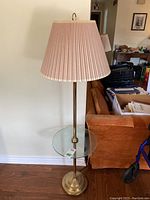 Full view of brass-toned metal floor lamp with integrated clear glass round table and pleated fabric lampshade.