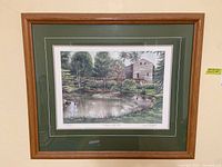 Framed print showing a pond and water mill with trees, double matted and behind glass in wood frame.