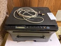 Samsung SCX-4300 monochrome multi-function laser printer in black and gray with USB and power cables on top, placed in a basement setting.