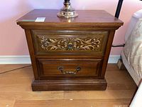 Photo of one Malcolm brand wood and wood laminate night stand showing two drawers, decorative carvings on the top drawer, metal handles on bottom drawer, medium brown wood finish.