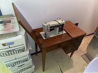 Three-drawer wooden sewing machine table with hinged top, shows surface wear and scratches, sewing machine mounted in center.