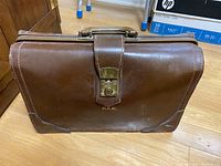 Front angle showing brown leather briefcase with top handle, gold-tone lock and R.E.K. initials.
