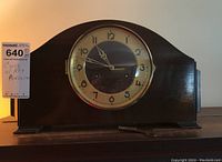 Front view of the vintage wooden mantel clock on a shelf showing the curved wooden case, brass bezel, and clock face with Arabic numerals and hands. Pendulum and key are placed in front.