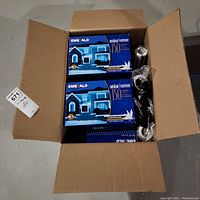 Opened cardboard box containing four visible boxes of Emerald outdoor Christmas lights and several paint rollers wrapped in plastic.