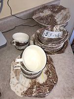 Stacked plates and coffee cups with saucers on countertop showing brown transferware village design