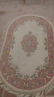 Full view of the oval wool rug showing floral design, pink border and fringe ends on carpeted floor