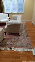 Photo showing the overall wool rug positioned under cream upholstered furniture, visible fringe on one edge, and hardwood floor beneath.