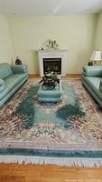 Full view of large wool rug with green background and floral border. Rug placed on hardwood floor between green leather sofas and a fireplace.