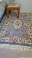 Light blue wool rug with floral central medallion under wooden side table, showing pattern and fringe.