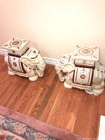 Pair of ceramic elephant side tables shown from an angle, showcasing both pieces side by side on hardwood flooring.