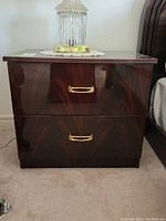 Front view of one nightstand showing two drawers, dark faux wood grain finish, and gold-tone hardware.