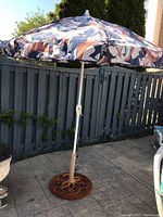 Full view of the patio umbrella with blue, orange, and white floral canopy and heavily rusted circular metal base on a patio.