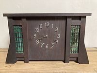 Front view of antique Mission style mantel clock showing dark wood case with two stained glass panels featuring green rounded rectangular pieces on either side of the clock face with metal numbers and missing clock hands.