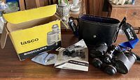 Photo showing the Tasco binoculars with their original yellow box, black carrying case, and instruction manual laid out on a wooden surface.