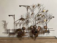 Front view showing the full metal wall art piece depicting a stylized park scene with trees, street lamps, benches, and umbrella-carrying figures