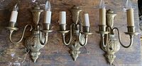 Three brass double wall sconces side by side on wooden surface. All have two arms with faux candle sleeves and bulbs. One arm is off the hook on one sconce.