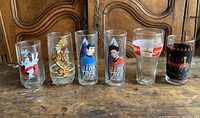 Six collectible drinking glasses including Star Trek, Ghostbusters, Winnie the Pooh, Coca-Cola, and 1984 Olympics themed glasses aligned on a wooden surface.