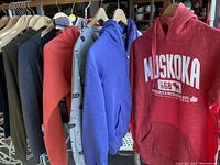 Photo showing multiple sweatshirts and pants hanging on a rack, including red Muskoka sweatshirt and blue zip-up hoodie.