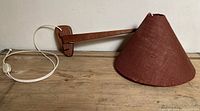 Full view of teak wall sconce arm with attached conical reddish-brown whipstitched shade, laying on a wood surface against a plain background.