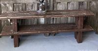 Photo showing full view of a rustic two-tier wooden bench with distressed paint and thick rectangular legs, situated against a wooden backdrop.