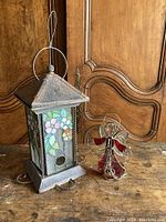 Photo showing stained glass lantern with floral design and metal frame alongside red stained glass angel candle holder on wood surface.
