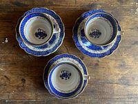 Three blue willow pattern porcelain tea cups paired with matching saucers, arranged on a wooden surface showing intricate blue and white design with gold trim.