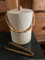 Full view of fabric-covered ice bucket with bamboo handle and matching bamboo tongs placed in front.