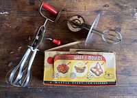 Photo showing two vintage hand mixers with red handles and chrome beaters alongside original boxed waffle mould set.