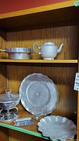 Shelf showing aluminum teapot, server with lid, small rectangular tray, and two decorative embossed trays