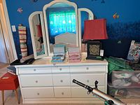 White wood dresser with six drawers and items on top including a lamp and storage boxes. Matching three-panel mirror positioned behind dresser.
