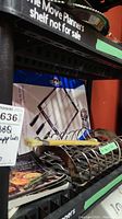 Photo of metal skewer rack, rib rack, and BBQ recipe cookbooks stacked on dark plastic shelf. Includes bamboo handled basting brush and assorted grill utensils.