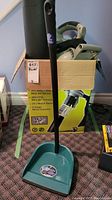 Photo showing the dustpan with black long handle standing upright in front of the cardboard box which contains the electric blower.